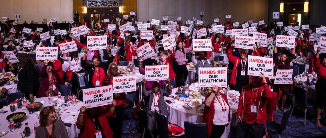 Nurses in large conference room holding signs that say "hands off our healthcare"