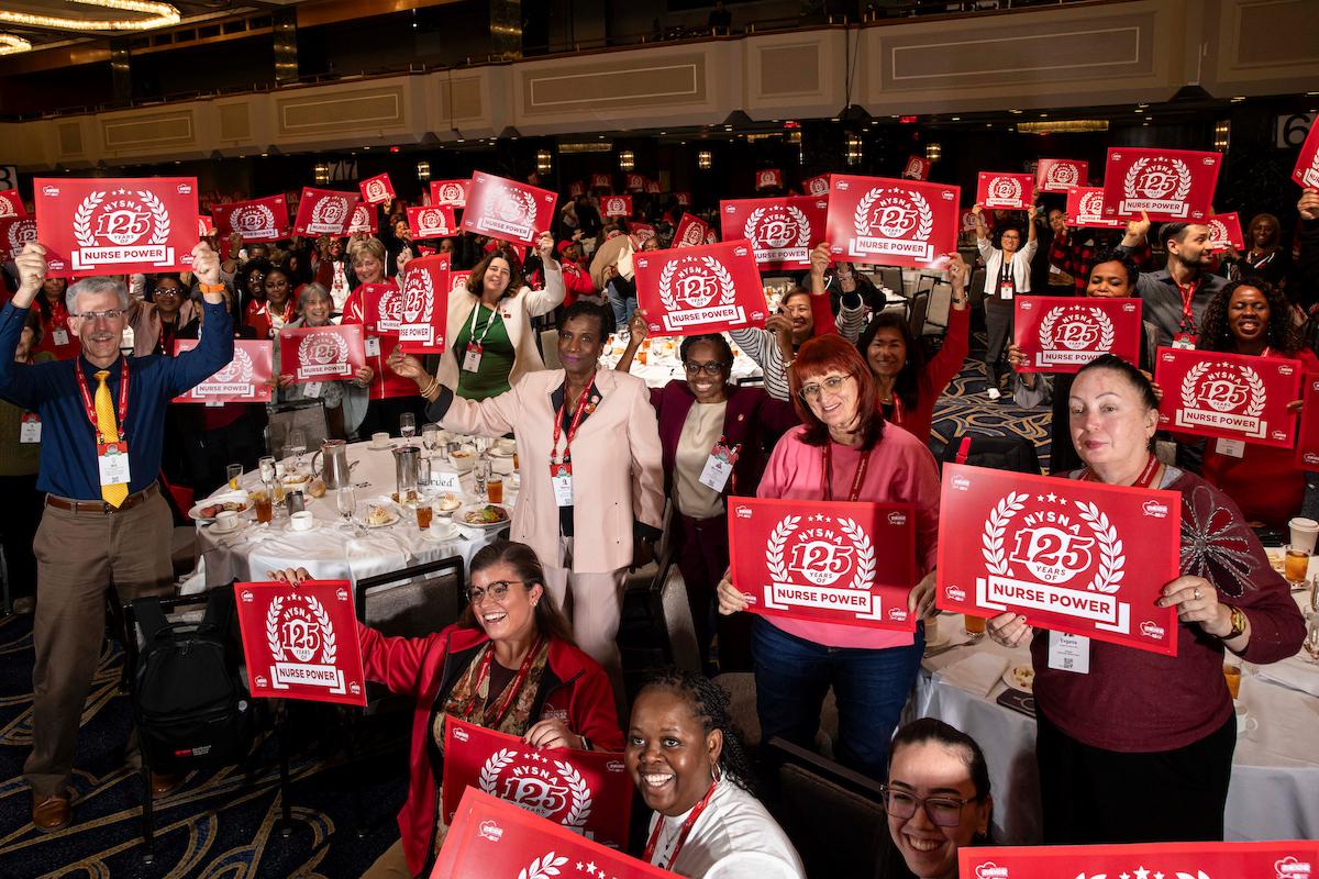 Crowd of nurses in a ballroom, holding signs celebrating NYSNA's 125th Anniversary