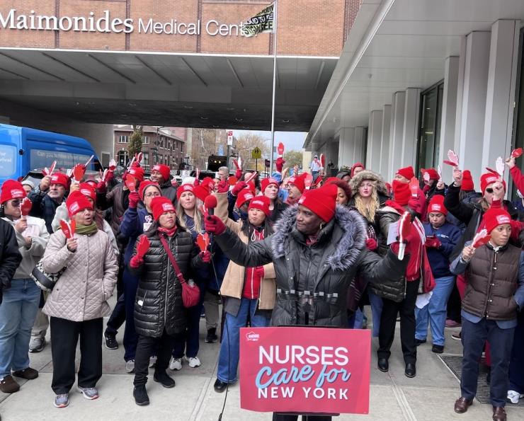 speaker at a microphone with a placard that says "Nurses care for New York" surrounded by nurses cheering