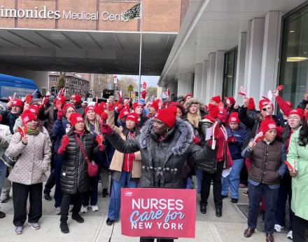 speaker at a microphone with a placard that says "Nurses care for New York" surrounded by nurses cheering